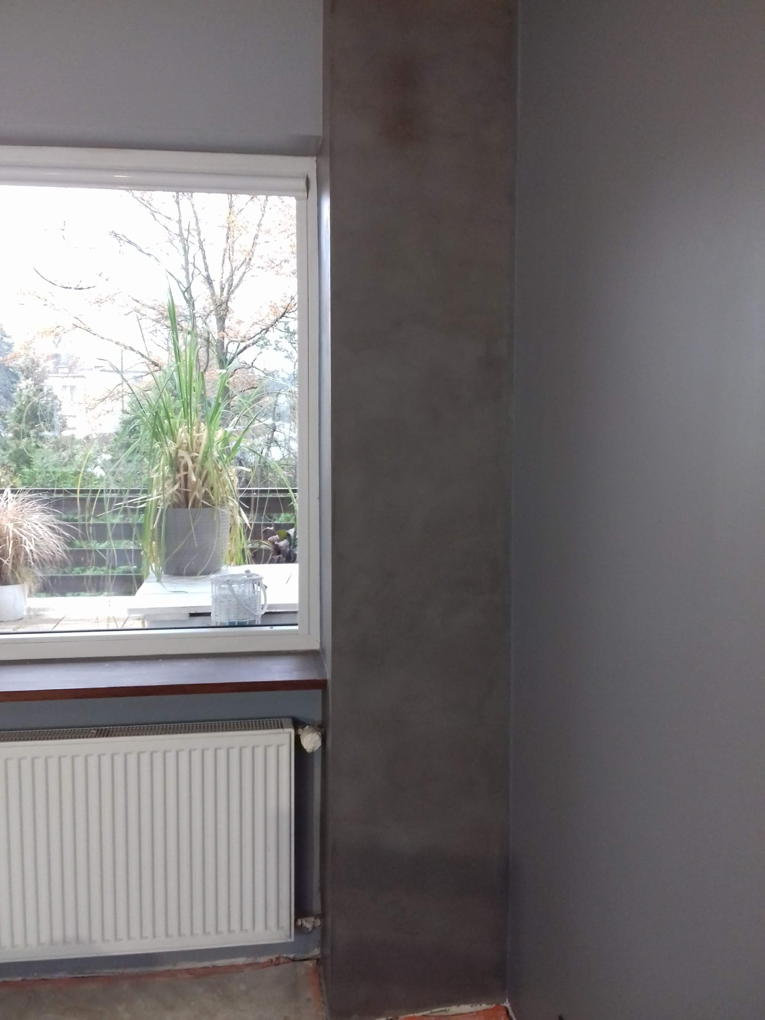 Wnętrze pokoju z oknem: ściana pomalowana na szaro z efektem dekoracyjnym w rogu, biały grzejnik pod oknem, za oknem widok na balkon i drzewa.