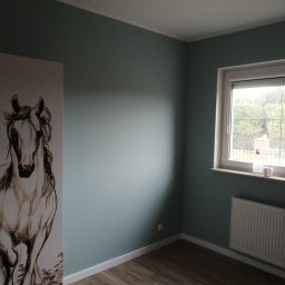 Łukasz Rogulski - Pomalowany na szaro-zielono pokój z drewnopodobną podłogą, grzejnikiem pod oknem i szafą z rysunkiem konia.