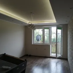 Pusty pokój po remoncie z drewnopodobną podłogą, ścianą z cegły dekoracyjnej, oświetleniem LED na suficie i otwartym balkonem z widokiem na zewnątrz.