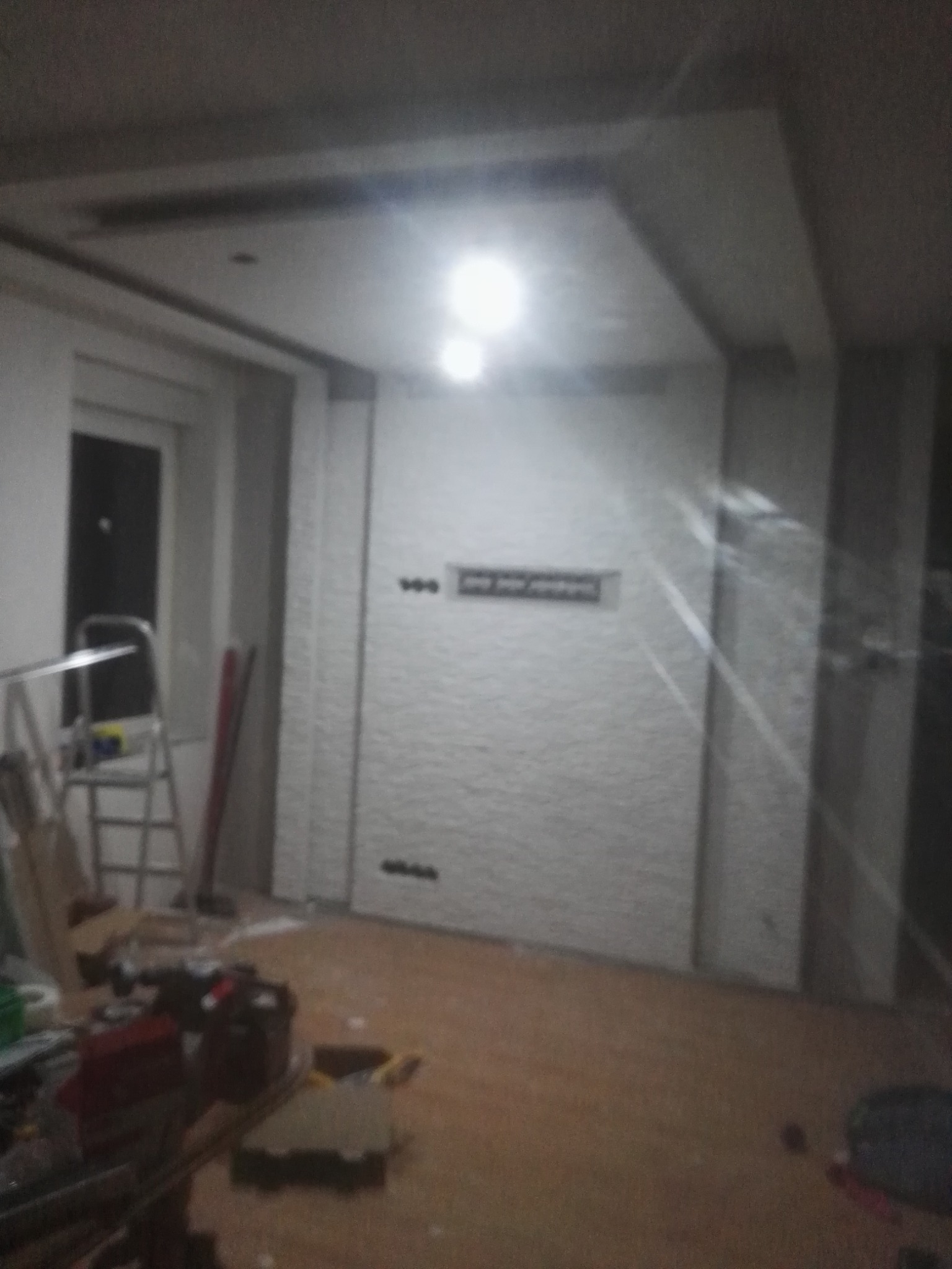 Wnętrze w trakcie remontu: ściana z fakturą cegły, widoczne puszki elektryczne, sufit podwieszany z oświetleniem, drabina i narzędzia malarskie na podłodze.