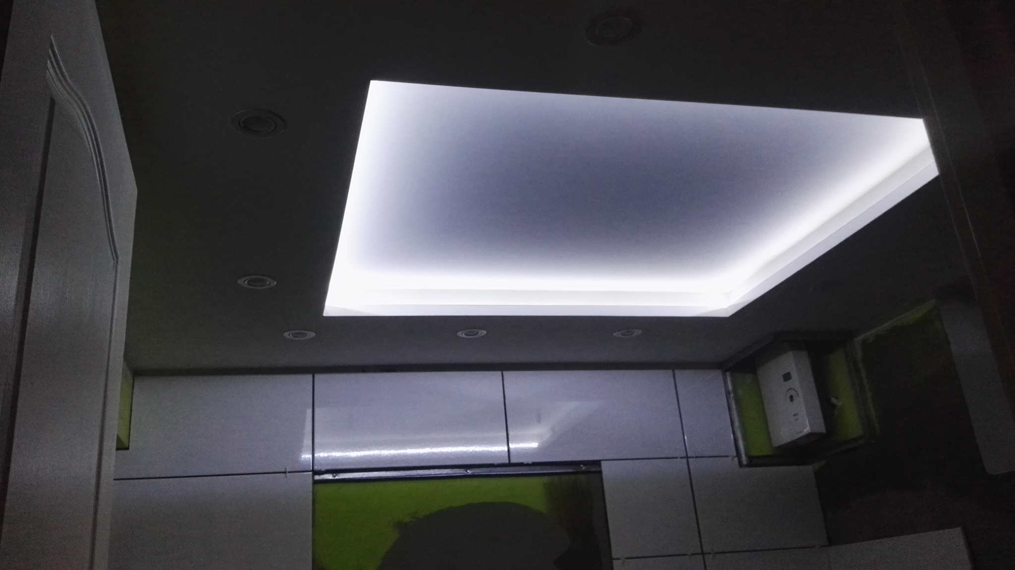 Nowoczesna kuchnia z podświetlanym sufitem w kształcie prostokąta i punktowymi lampami, szafki z połyskiem, widoczny fragment drzwi i zabudowany piec.