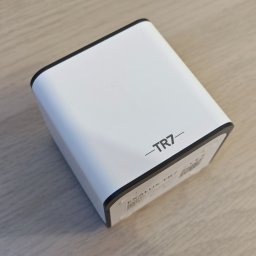 Xdom inteligentne domy - Biała centralka inteligentnego domu TR7 z czarnymi krawędziami, leżąca na jasnym drewnianym blacie, widok z góry pod kątem.