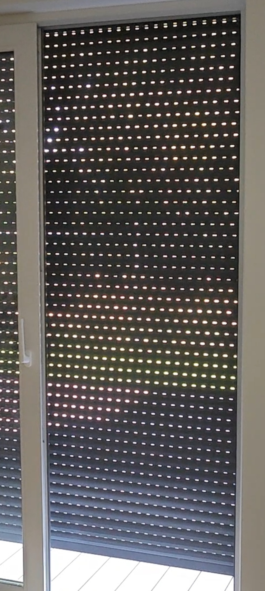 Częściowo opuszczona, perforowana roleta zewnętrzna w oknie, widoczne światło dzienne wpadające przez otwory.