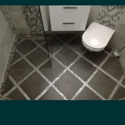 Wykończona łazienka z ciemną podłogą w kratkę z błyszczącymi fugami, jasnymi płytkami na ścianach, białą szafką pod umywalką i toaletą.