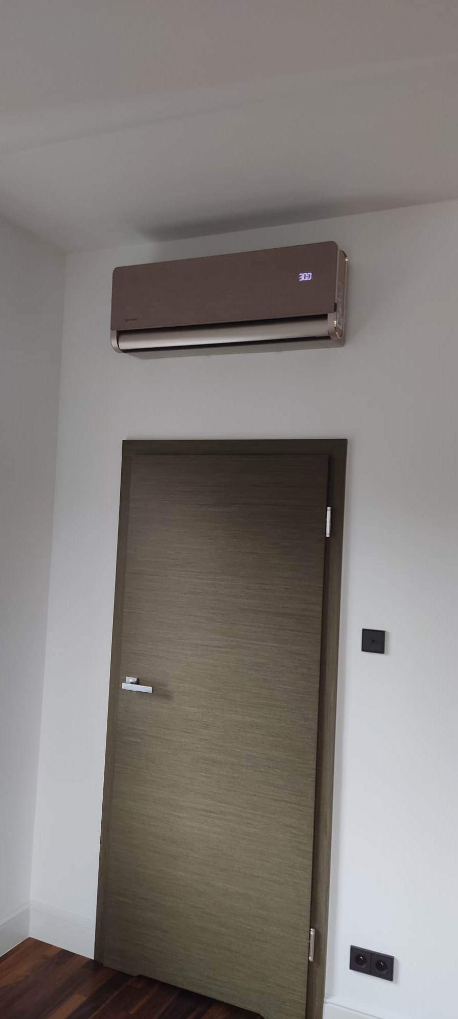 Beżowy klimatyzator ścienny z wyświetlaczem, zamontowany nad drewnianymi drzwiami w nowoczesnym wnętrzu o białych ścianach i ciemnej podłodze.