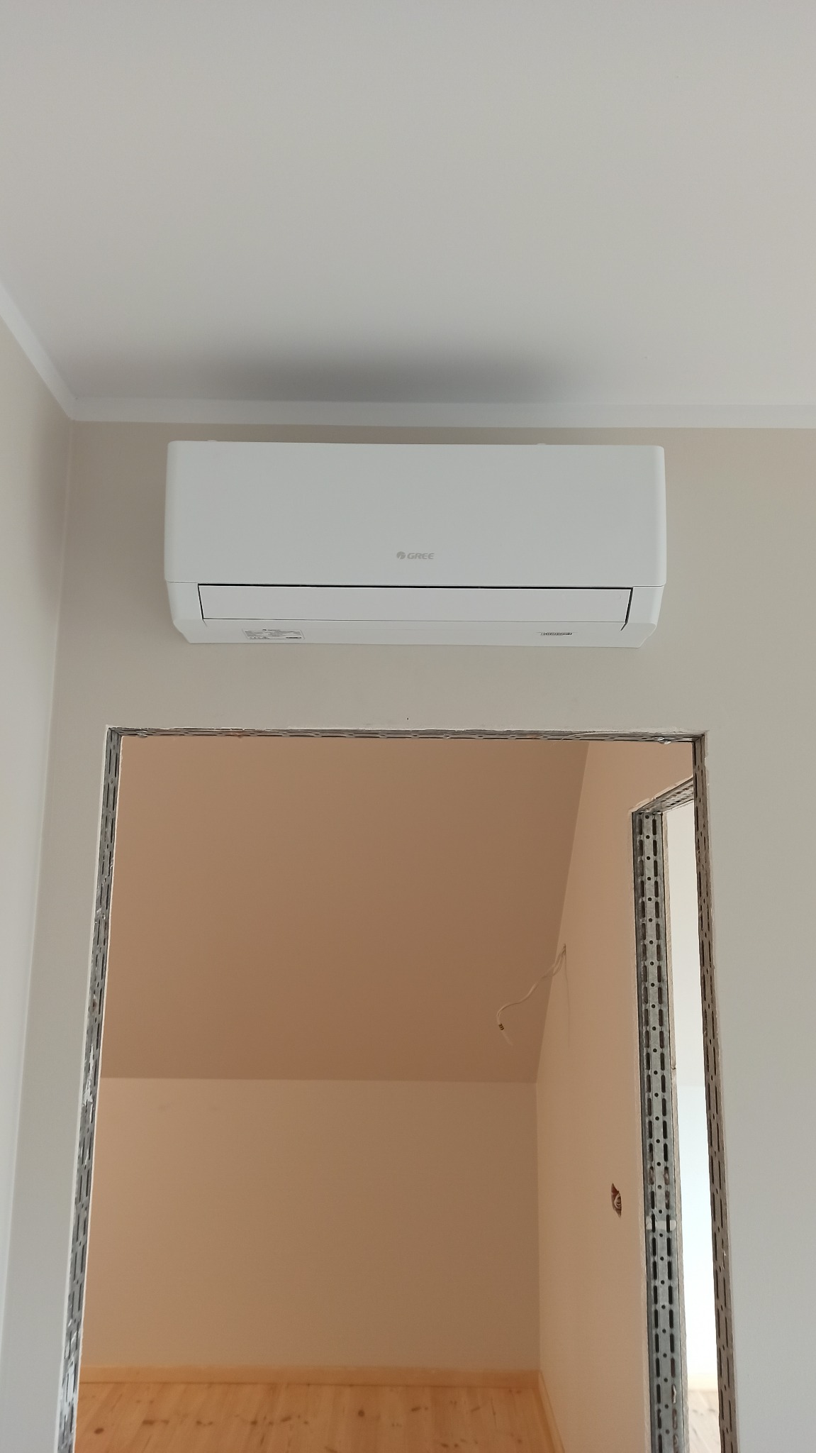 Biały klimatyzator marki Gree zamontowany na ścianie nad surowym otworem drzwiowym w jasnym pomieszczeniu w trakcie remontu.