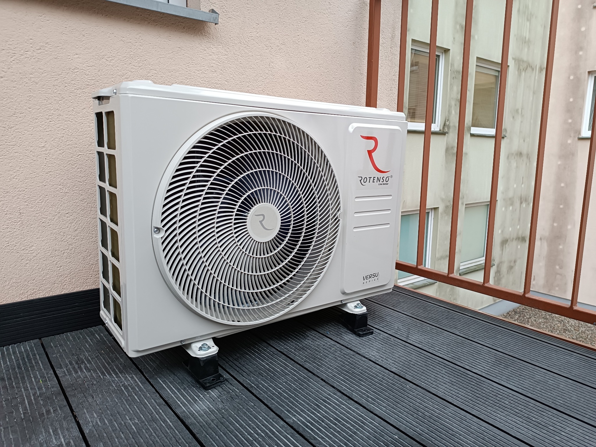 Biała jednostka zewnętrzna klimatyzacji Rotenso Versu Series zamontowana na czarnym tarasie balkonowym, z widocznymi budynkami w tle.