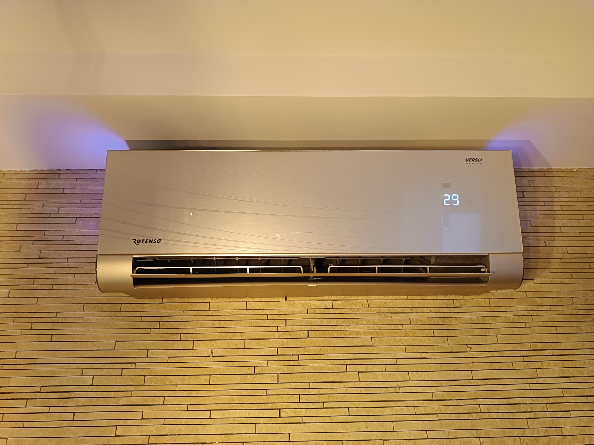 Nowoczesny klimatyzator Rotenso Versu Series zamontowany na ścianie z dekoracyjnej cegły, wyświetlający temperaturę 29 stopni.