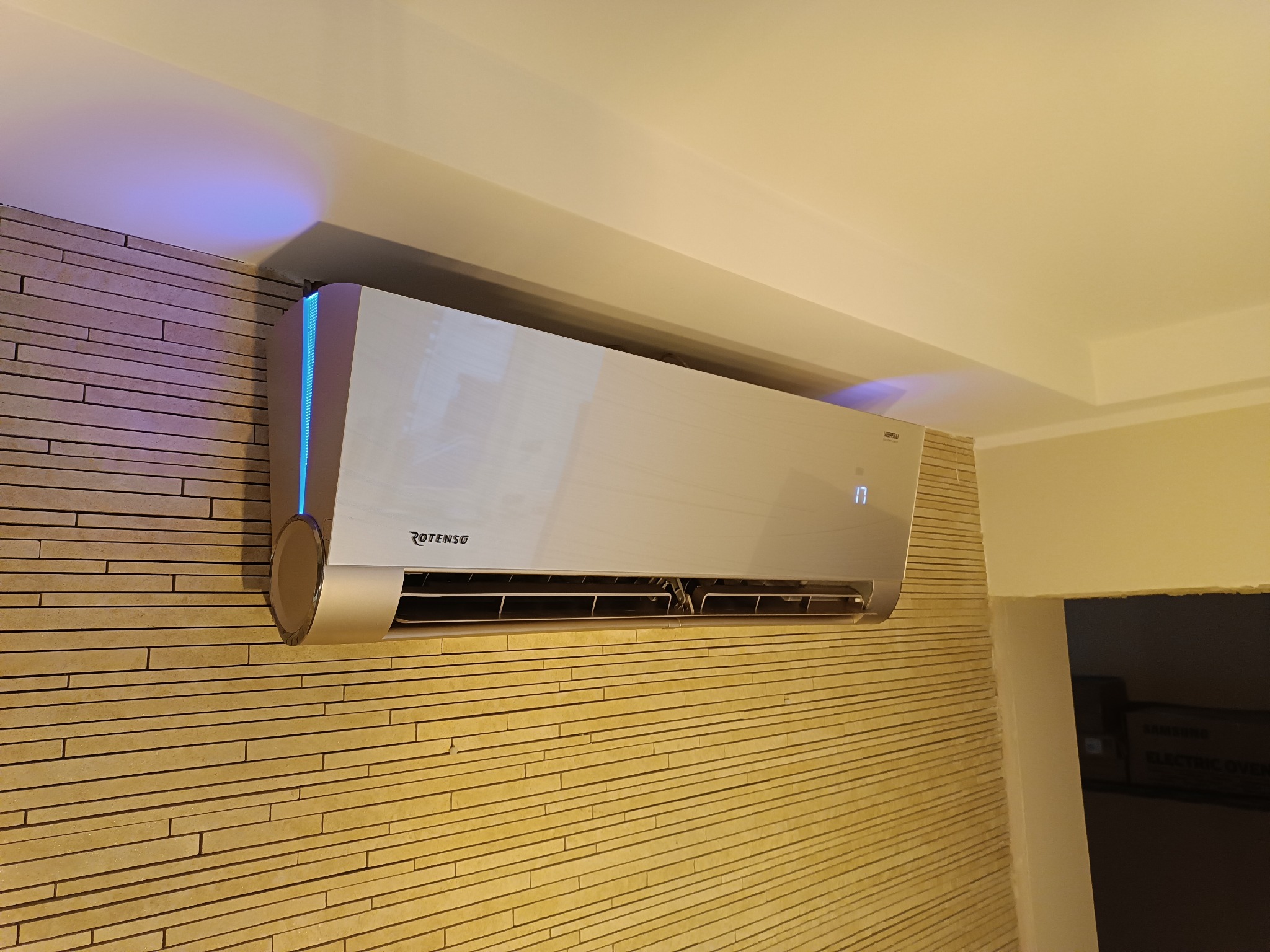 Zamontowany klimatyzator marki Rotenso na ścianie wyłożonej dekoracyjną cegłą, z widocznym niebieskim podświetleniem bocznym i wyświetlaczem temperatury.