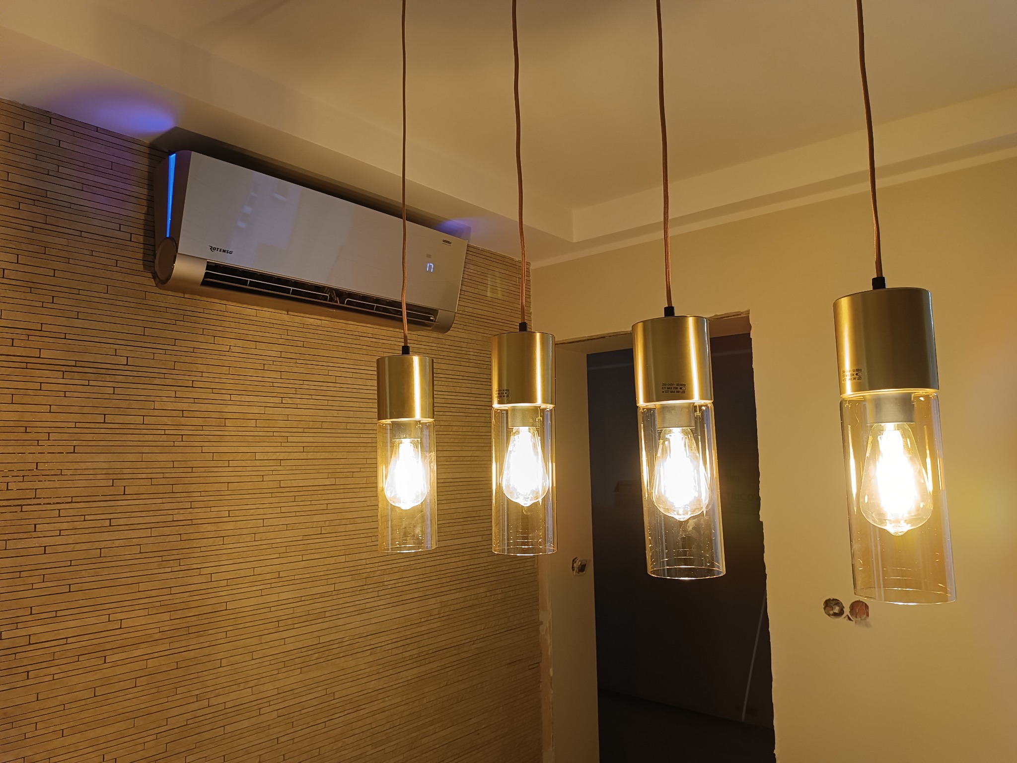 Nowoczesny klimatyzator ścienny Rotenso z niebieskim podświetleniem LED, zamontowany nad czterema złotymi lampami wiszącymi z przezroczystymi kloszami, na tle ściany z dekoracyjną okładziną...