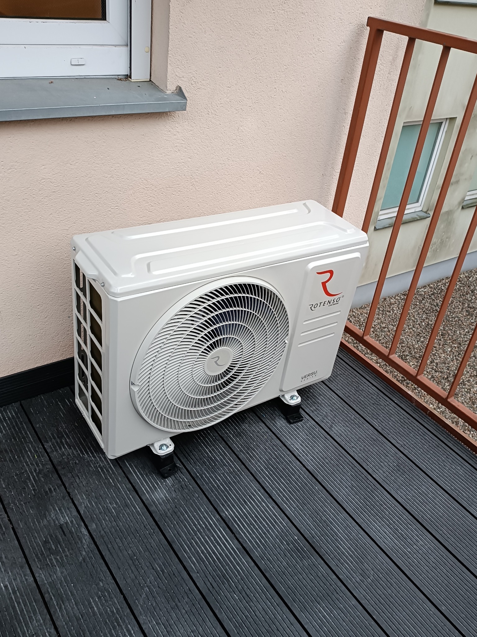 Zewnętrzna jednostka klimatyzacji Rotenso Versu Series zamontowana na czarnym tarasie, widok z góry, obok brązowa barierka.