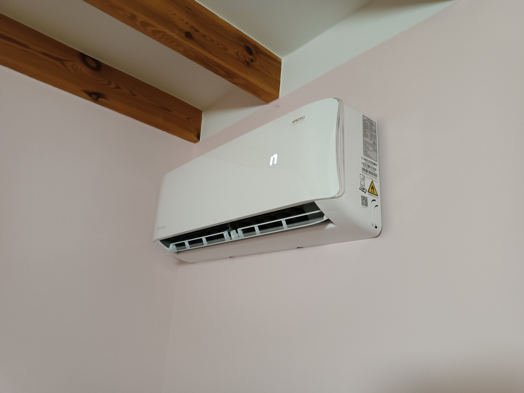 Biała jednostka klimatyzatora IMOTO zamontowana na jasnoróżowej ścianie w pomieszczeniu z drewnianymi belkami stropowymi.