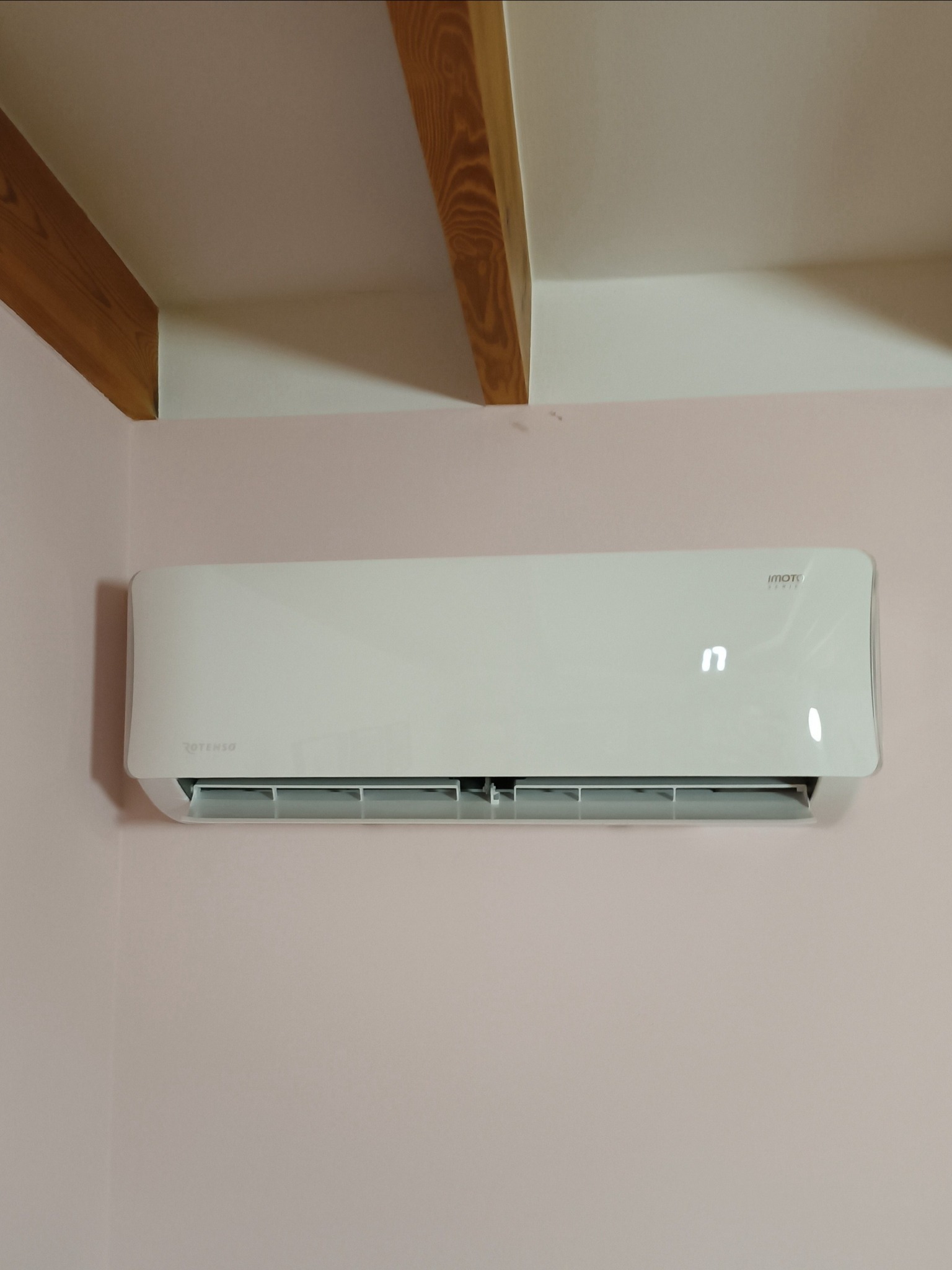 Zamontowany na różowej ścianie klimatyzator Rotenso Imoto z wyświetlaczem temperatury, widoczny fragment drewnianej belki stropowej.