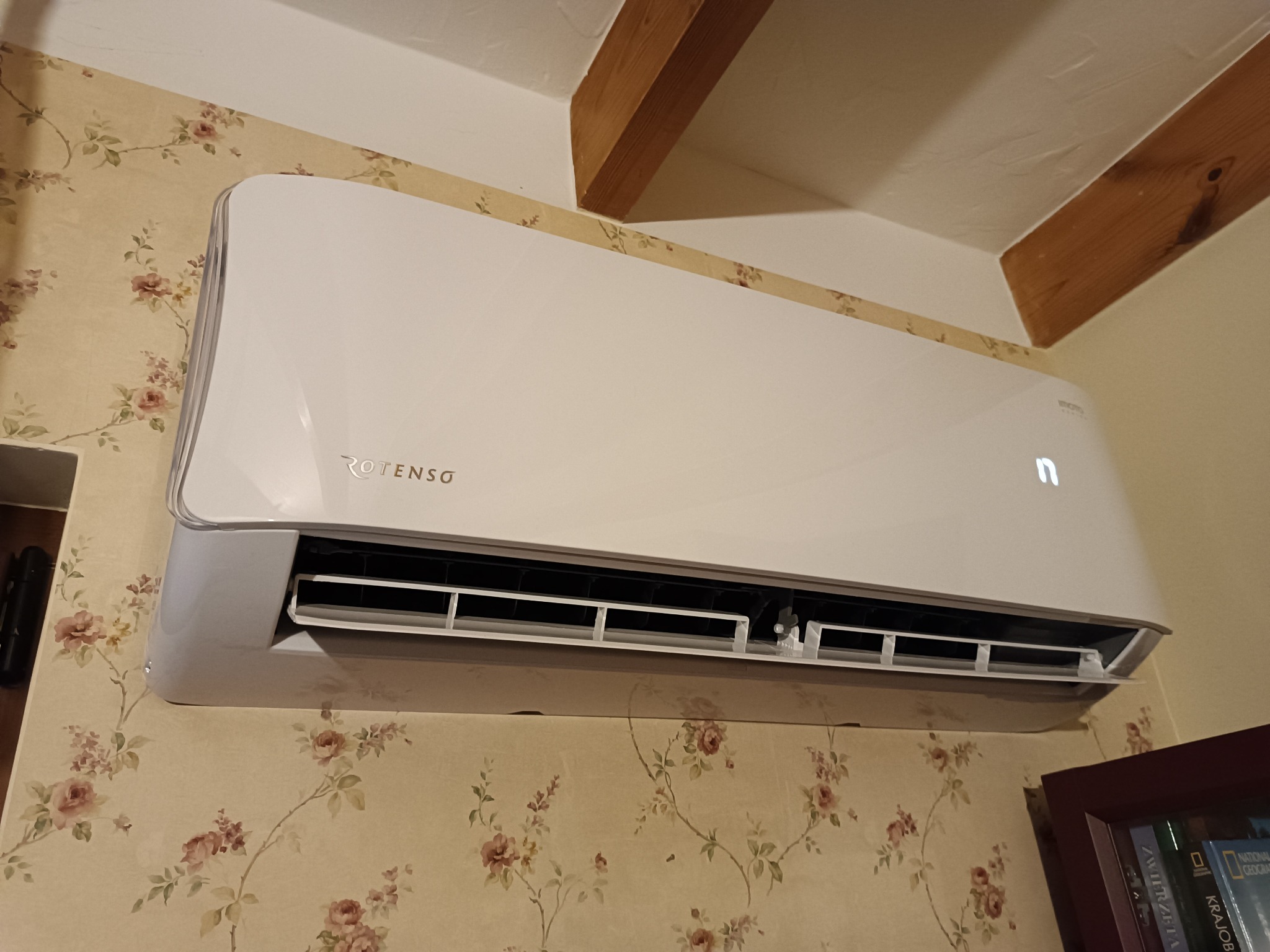 Biały klimatyzator ROTENSO zamontowany na ścianie z tapetą w kwiaty, widoczne drewniane belki stropowe.