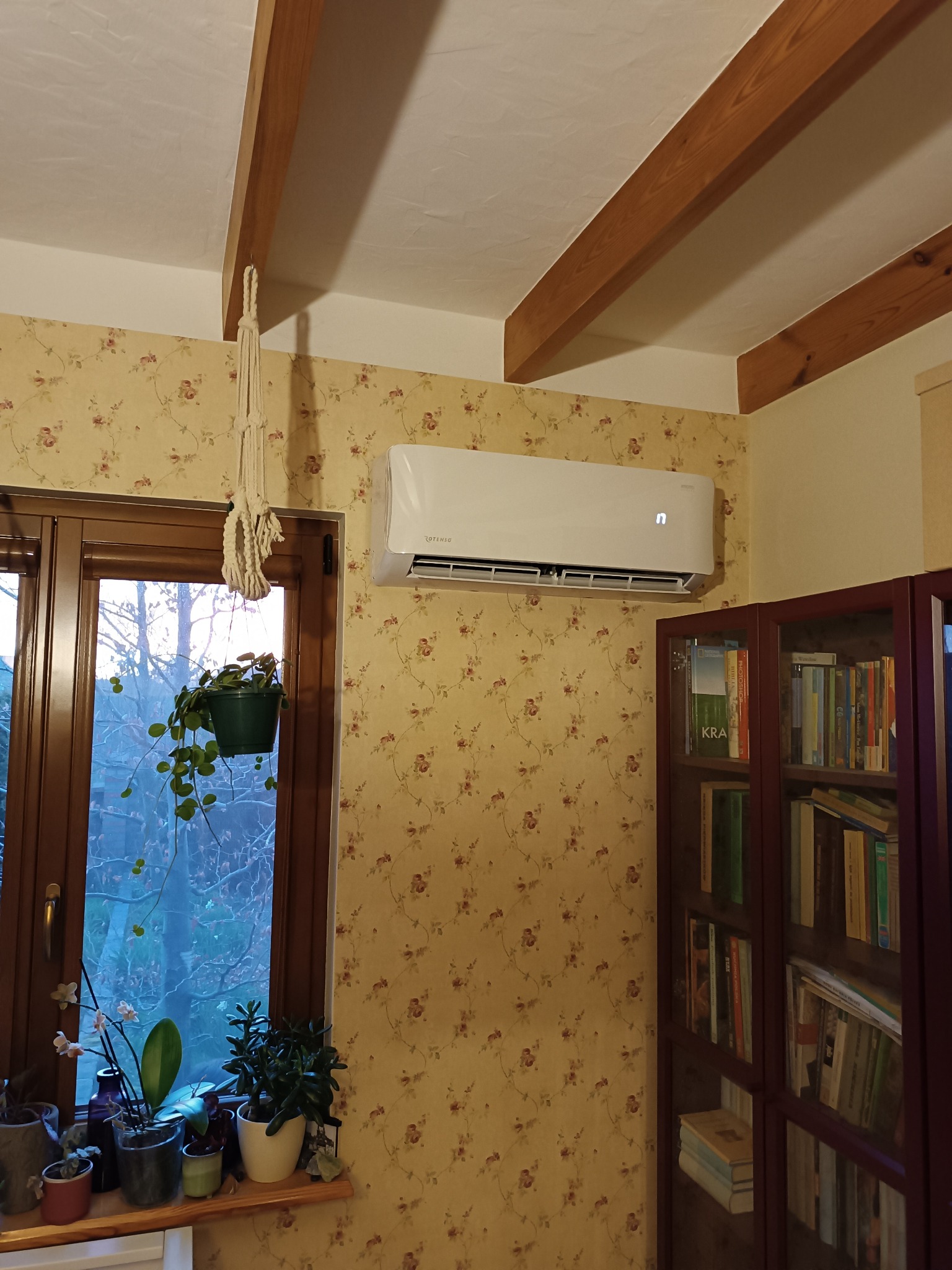 Biały klimatyzator Rotenso zamontowany na ścianie z tapetą w kwiaty, widok na drewniane belki stropowe, rośliny doniczkowe na parapecie oraz regał z książkami w tle.