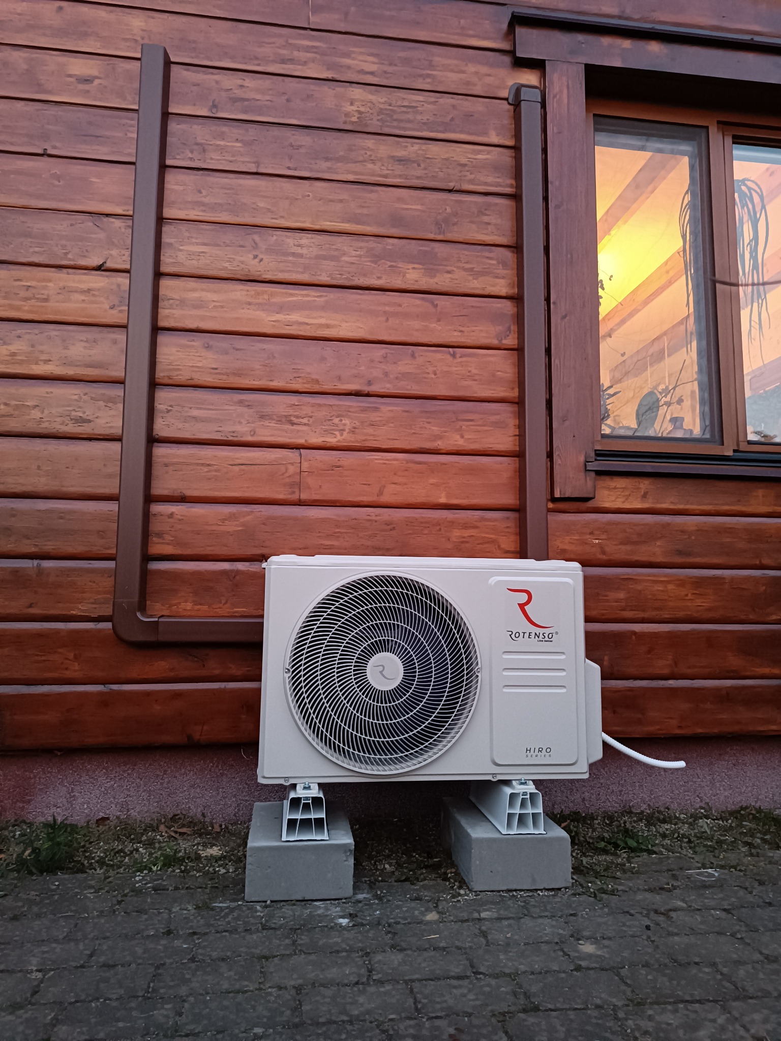 Zamontowana jednostka zewnętrzna klimatyzacji Rotenso Hiro na betonowych podstawach przy drewnianej ścianie domu z widocznym oknem, odbijającym zachodzące słońce.