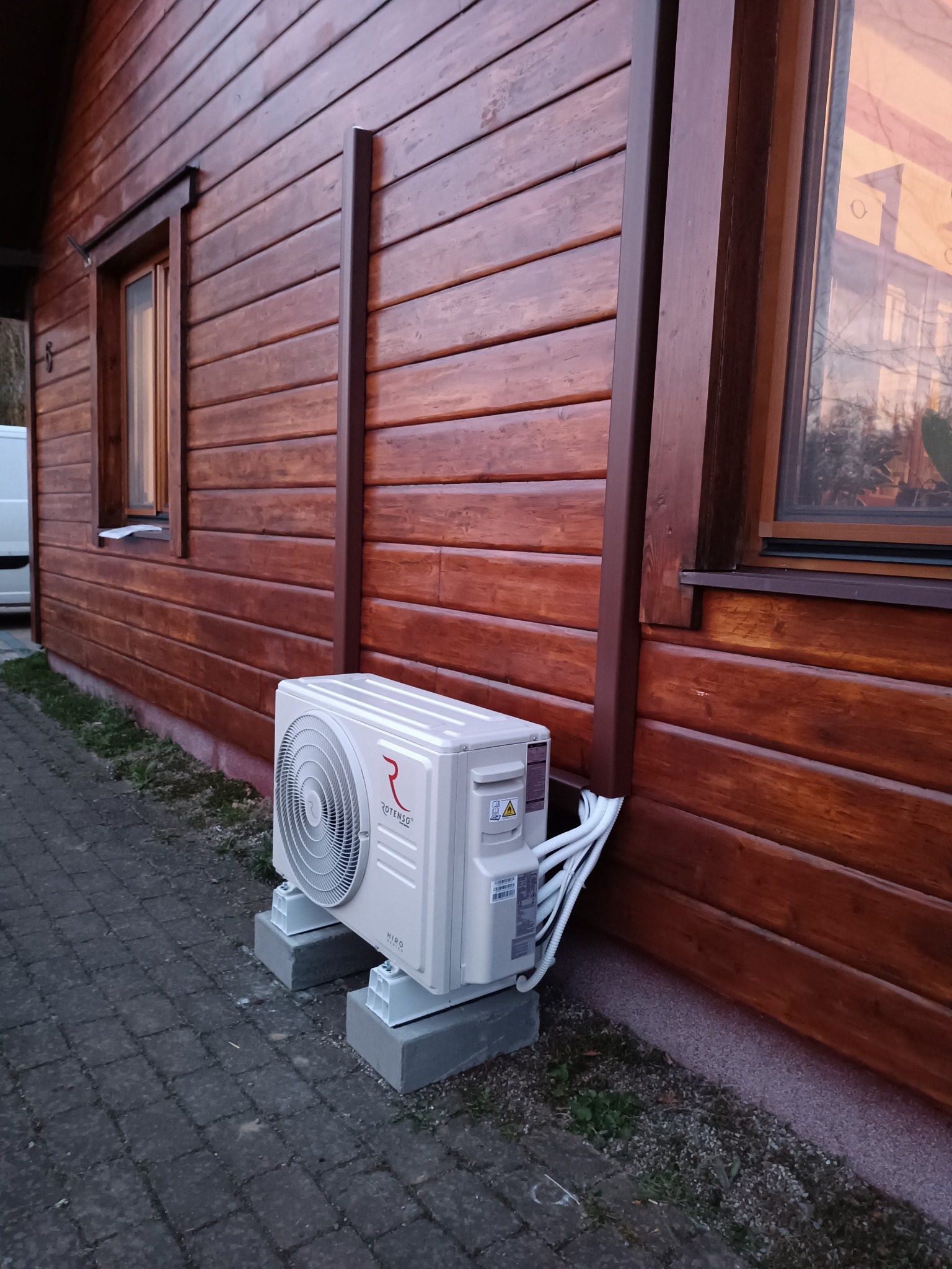 Jednostka zewnętrzna klimatyzacji Rotenso zamontowana na betonowych bloczkach przy drewnianym domu z widocznymi przewodami w peszlu.