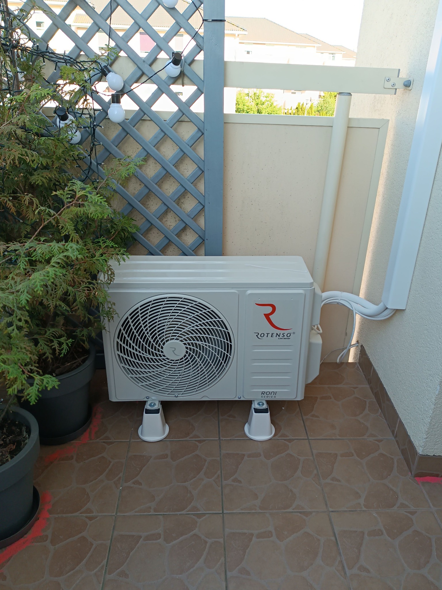 Zewnętrzna jednostka klimatyzacji Rotenso zamontowana na balkonie, widoczne przewody prowadzone wzdłuż ściany, obok donica z tujami i dekoracyjne oświetlenie.