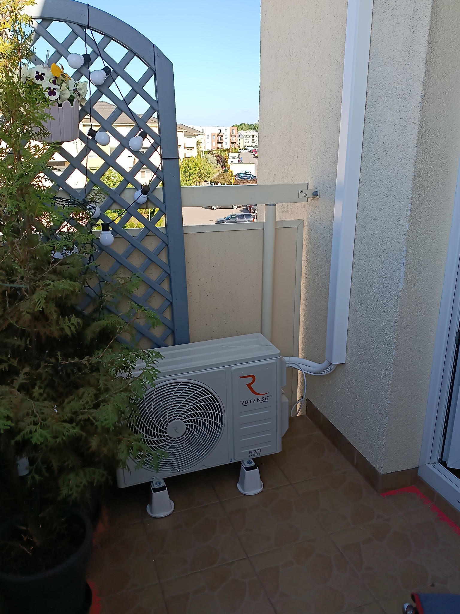 Jednostka zewnętrzna klimatyzacji marki Rotenso zamontowana na balkonie, z widocznymi przewodami w białej maskownicy, na tle zabudowy miejskiej.