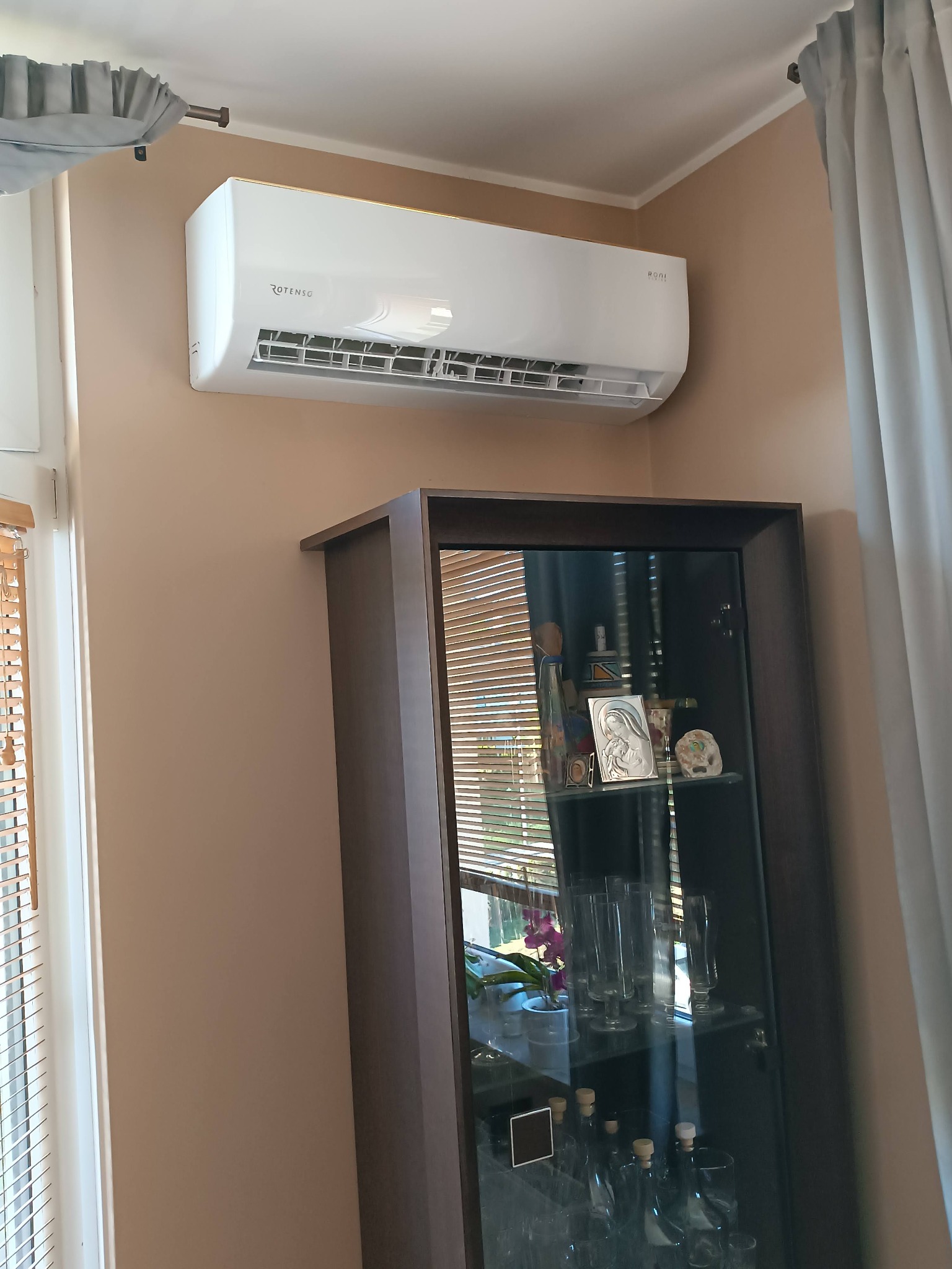 Biała klimatyzacja marki Rotenso zamontowana na beżowej ścianie nad ciemnobrązową witryną z przeszklonymi drzwiami, widoczne zasłony i żaluzje.