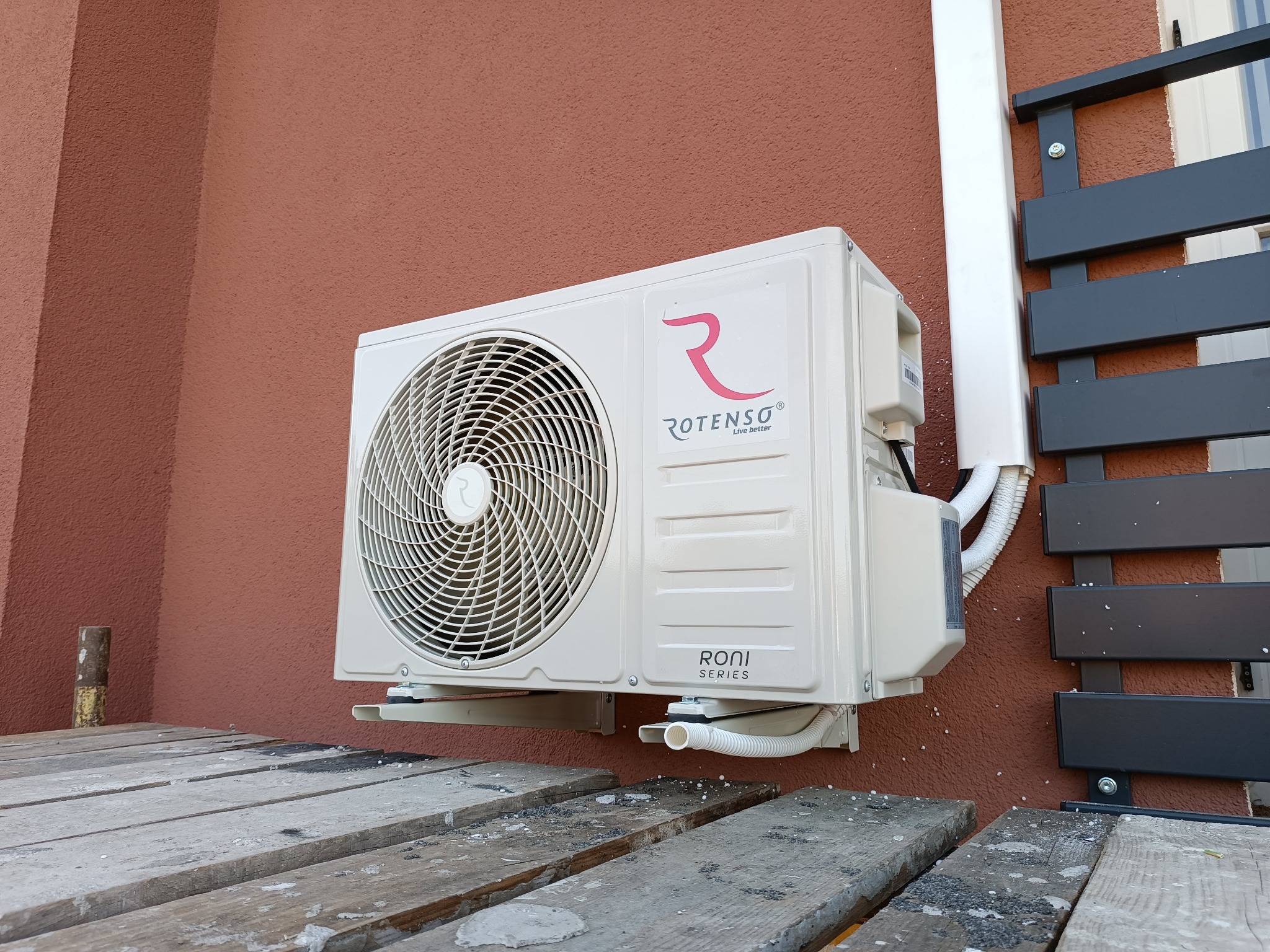 Zamontowana jednostka zewnętrzna klimatyzacji Rotenso Roni Series na brązowej ścianie, widoczna instalacja odprowadzająca skropliny, zarys drewnianego podestu w dolnej części kadru.