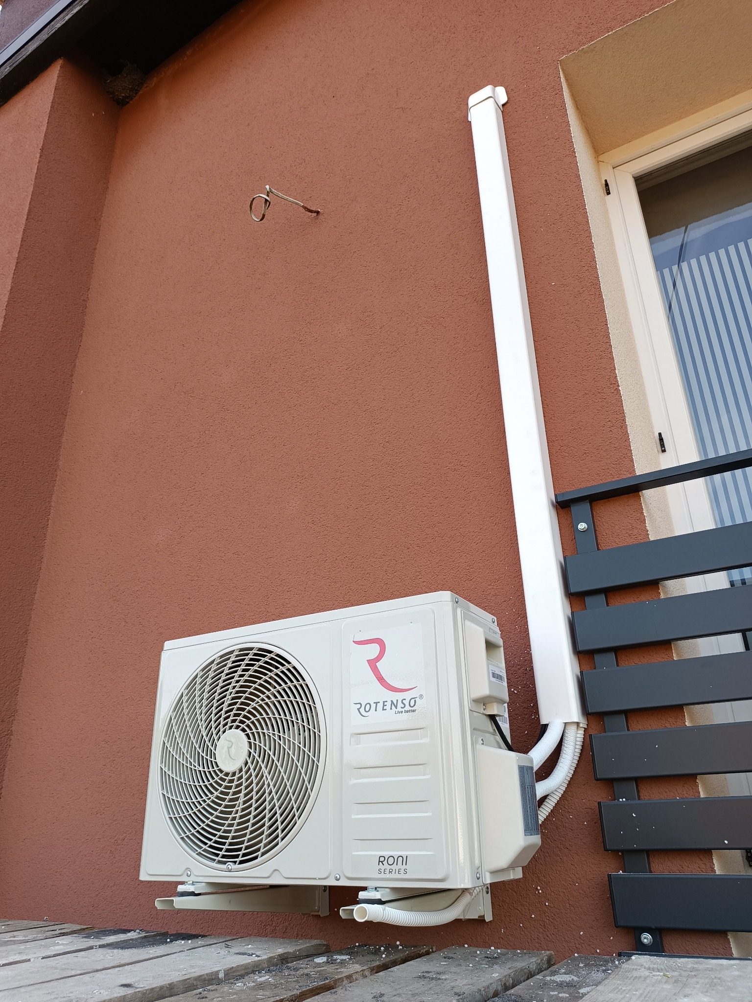 Zamontowana jednostka zewnętrzna klimatyzatora Rotenso Roni Series na brązowej elewacji budynku, widoczna instalacja w białej maskownicy, fragment okna i czarnego balustrady.