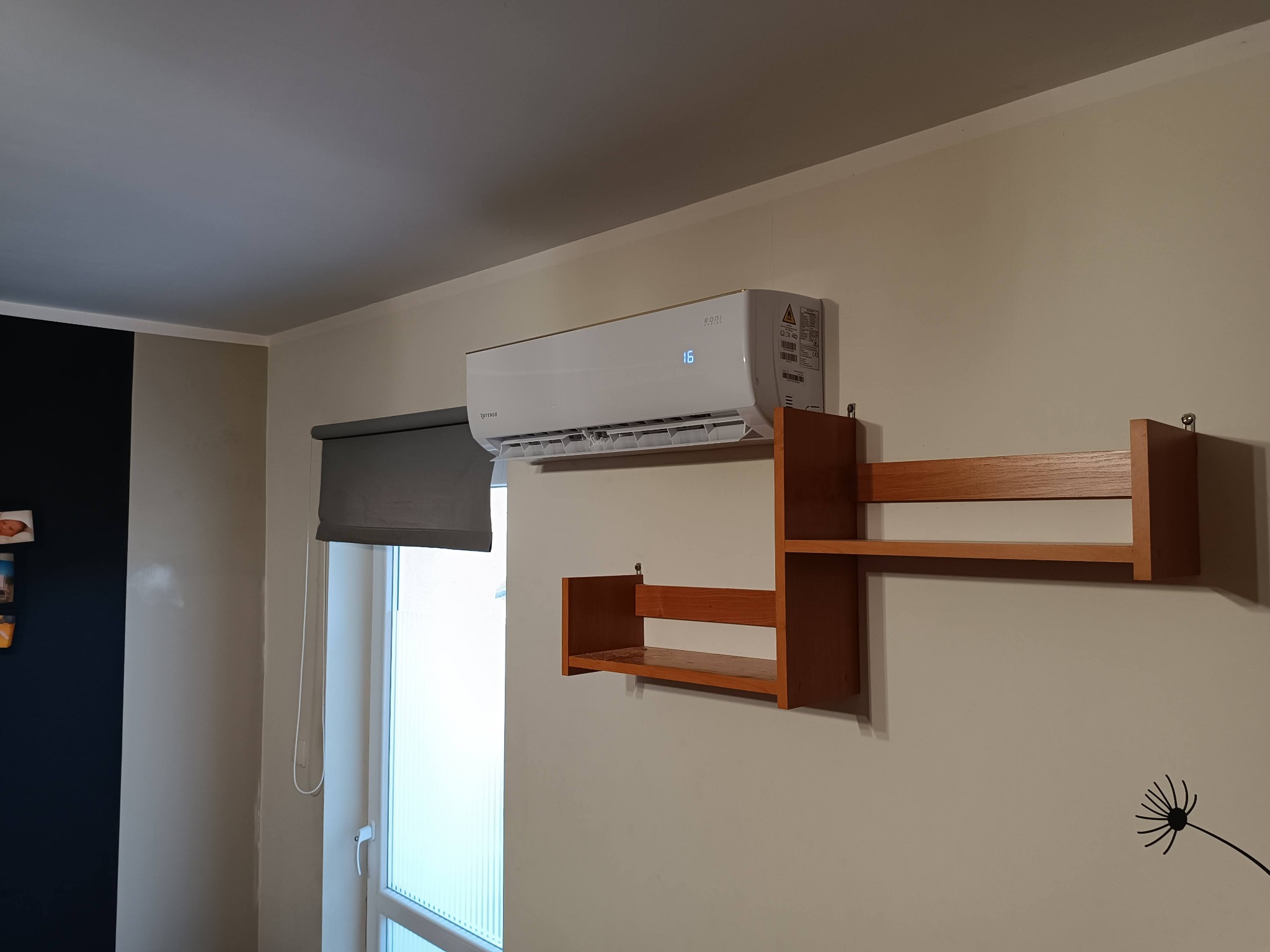 Biała jednostka klimatyzacji Rotenso zamontowana na ścianie nad brązową półką, widoczny wyświetlacz LCD wskazujący temperaturę 16 stopni Celsjusza, roleta okienna częściowo zasłonięta.