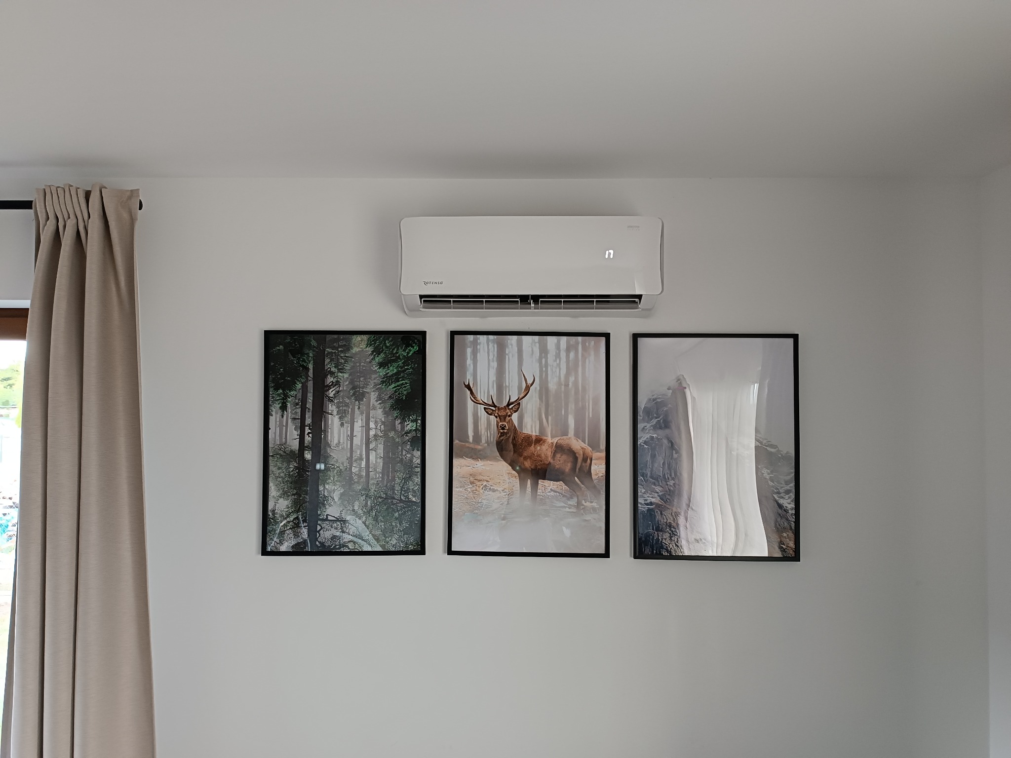 Biały klimatyzator wiszący na ścianie nad trzema obrazami w czarnych ramkach, obok zasłony.