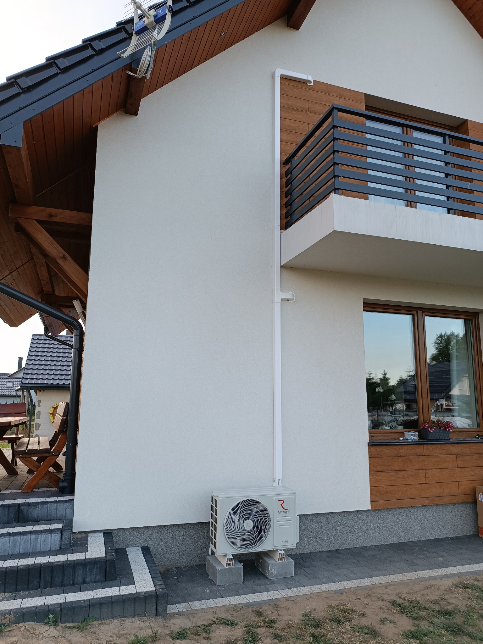 Zewnętrzna jednostka klimatyzacji Rotenso zamontowana na betonowych bloczkach przy ścianie domu w Rumi, z widoczną białą rurą prowadzącą wzdłuż elewacji i fragmentem schodów wejściowych.