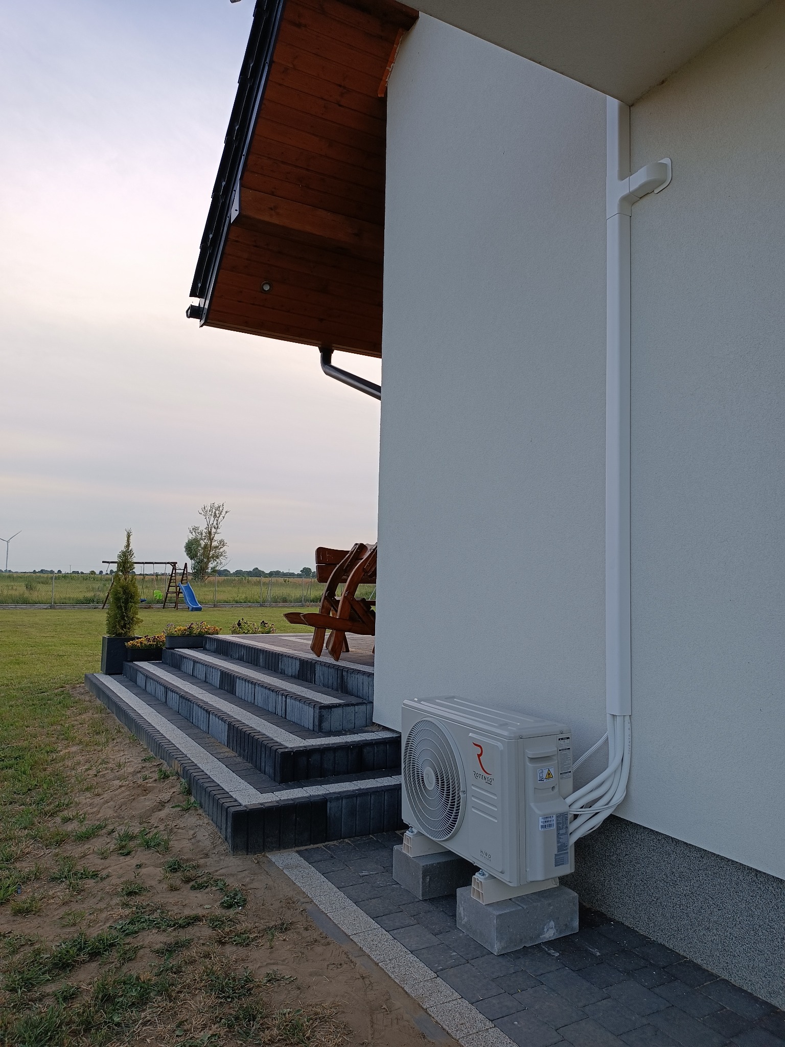 Zewnętrzna jednostka klimatyzacji zamontowana na betonowych bloczkach przy ścianie domu z widocznymi przewodami schowanymi w listwie PCV, w tle schody z kostki brukowej i trawnik.