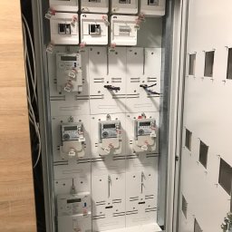 Szteker Elektryka - Otwarta szafa rozdzielcza z zamontowanymi licznikami energii elektrycznej i zabezpieczeniami, widoczne przewody i oznaczenia numeracyjne.