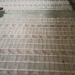 ElektroComplex - Rozłożona mata grzewcza Thermoval z czerwonymi przewodami na szarej, surowej posadzce przygotowana do wylewki.