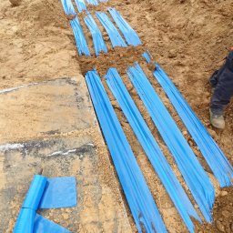 ElektroComplex - Wykop z ułożoną niebieską folią ochronną, prawdopodobnie zabezpieczającą kable elektryczne, widok z góry na brązową ziemię i fragment nogi w roboczym obuwiu.