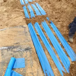 Wykop z ułożoną niebieską folią ochronną, prawdopodobnie zabezpieczającą kable elektryczne, widok z góry na brązową ziemię i fragment nogi w roboczym obuwiu.