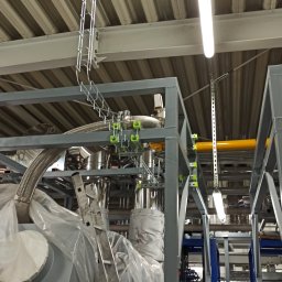 ElektroComplex - Widok na fragment hali produkcyjnej z instalacją przemysłową w trakcie montażu, z widocznymi elementami konstrukcji stalowej, rurami, przewodami w korytach kablowych oraz drabiną opartą...