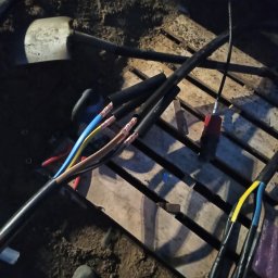 ElektroComplex - Fragment wykopu z ułożonymi przewodami elektrycznymi w czarnej izolacji, z widocznymi końcówkami kabli w kolorach niebieskim, żółtym i brązowym, przygotowanymi do połączenia, obok leży szpadel...
