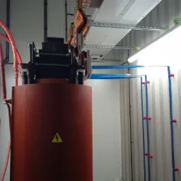 Brązowy transformator z żółtym znakiem ostrzegawczym na obudowie, widoczne miedziane przewody, szyny montażowe i oświetlenie w pomieszczeniu technicznym.