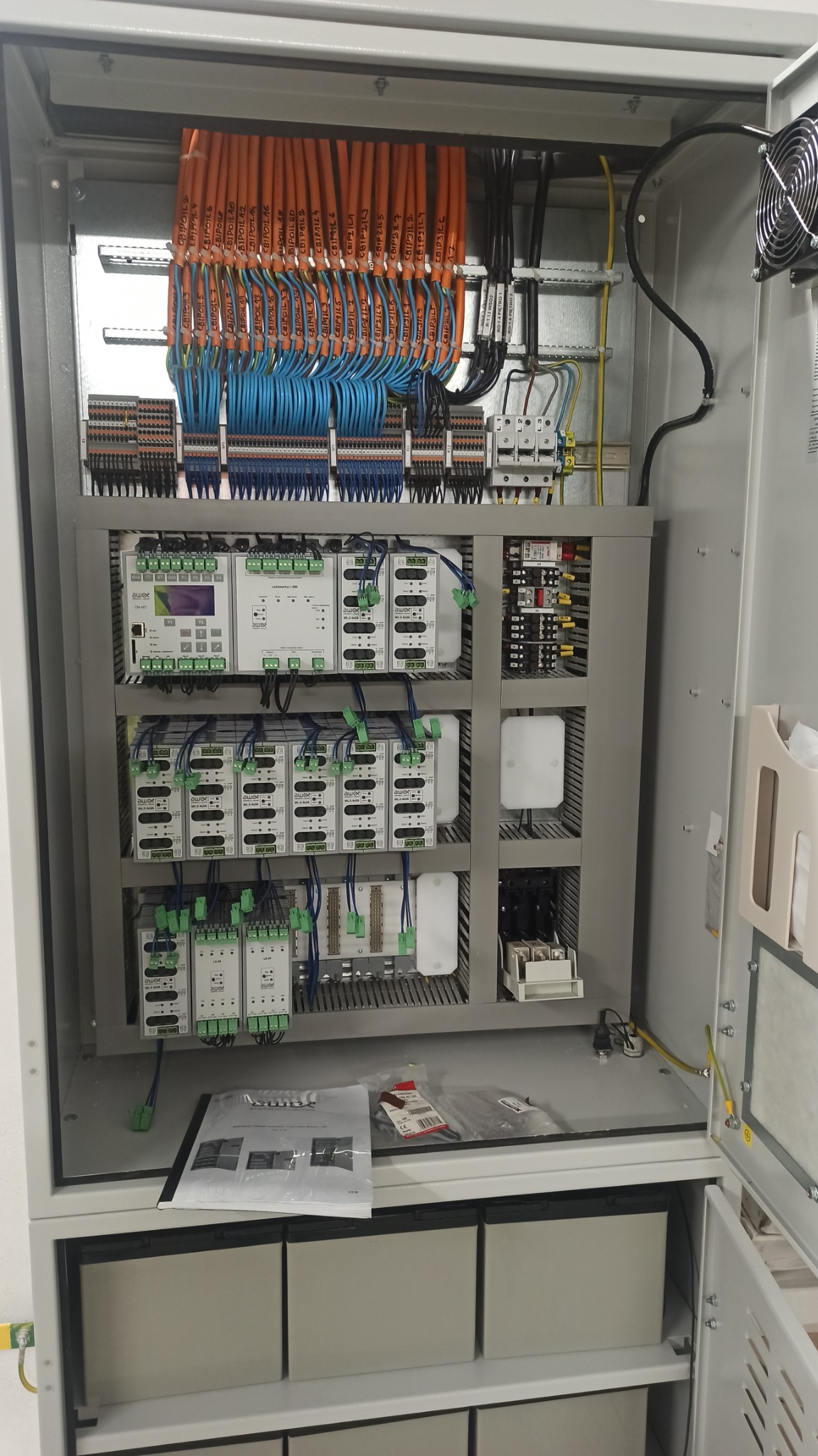 Otwarta szafa sterownicza z widocznymi modułami sterującymi, licznymi przewodami oznaczonymi etykietami, listwami zaciskowymi, zasilaczami i wentylatorem chłodzącym. Na półce leżą dokumenty...