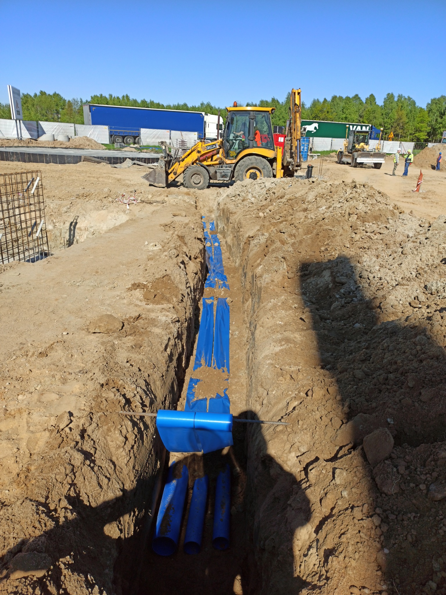 Wykop z ułożonymi niebieskimi rurami kanalizacyjnymi, zabezpieczonymi folią, na tle placu budowy z koparką i ciężarówkami w oddali.