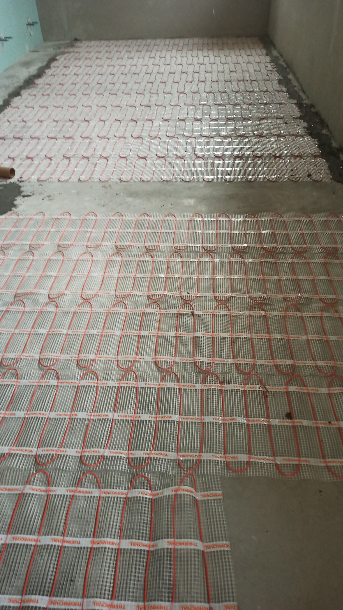 Rozłożona mata grzewcza Thermoval z czerwonymi przewodami na szarej, surowej posadzce przygotowana do wylewki.