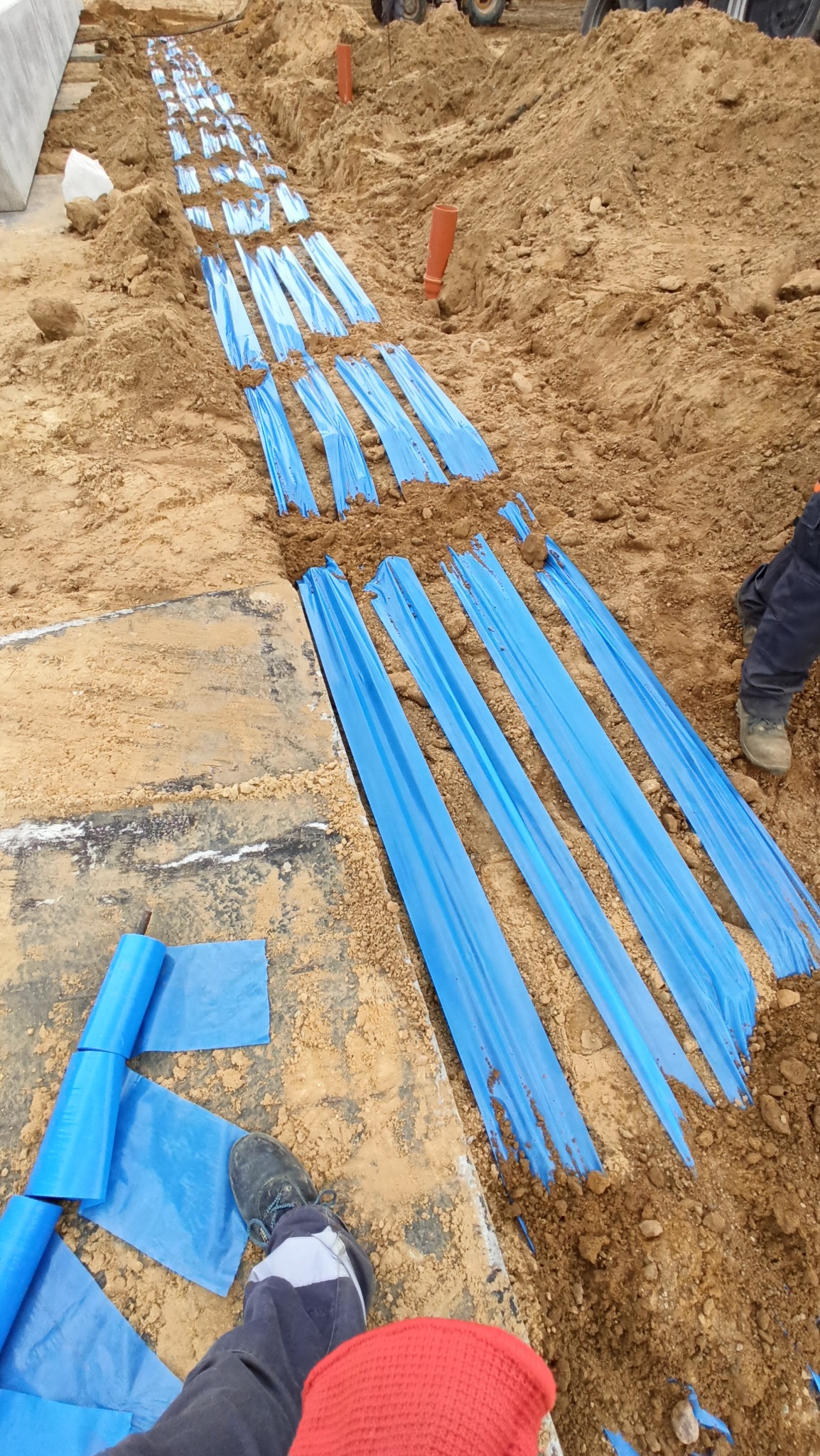 Wykop z ułożoną niebieską folią ochronną, prawdopodobnie zabezpieczającą kable elektryczne, widok z góry na brązową ziemię i fragment nogi w roboczym obuwiu.