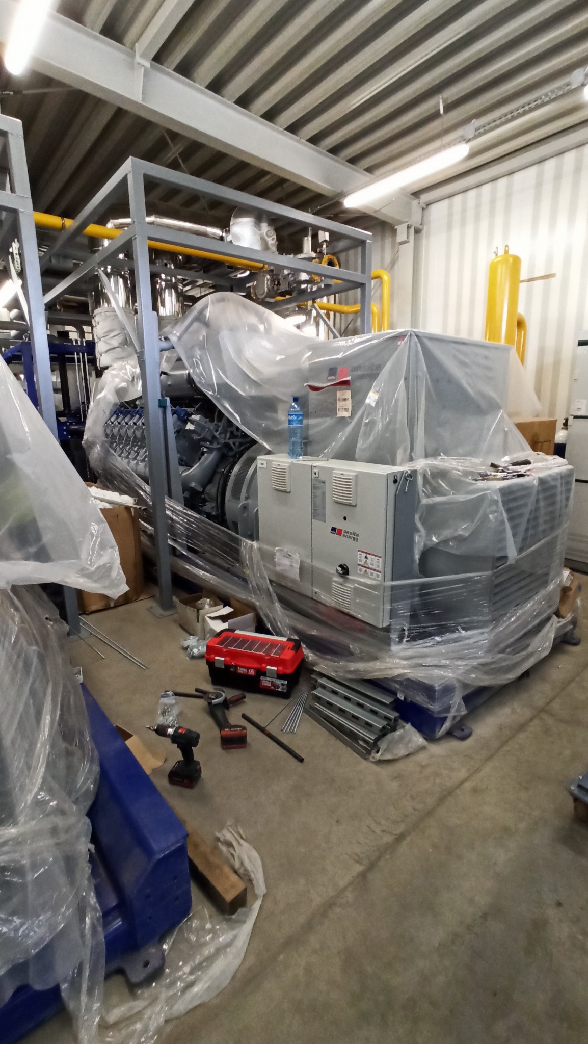 Agregat prądotwórczy w folii ochronnej, z widoczną skrzynką narzędziową i butelką wody, w hali z metalową konstrukcją stropu.