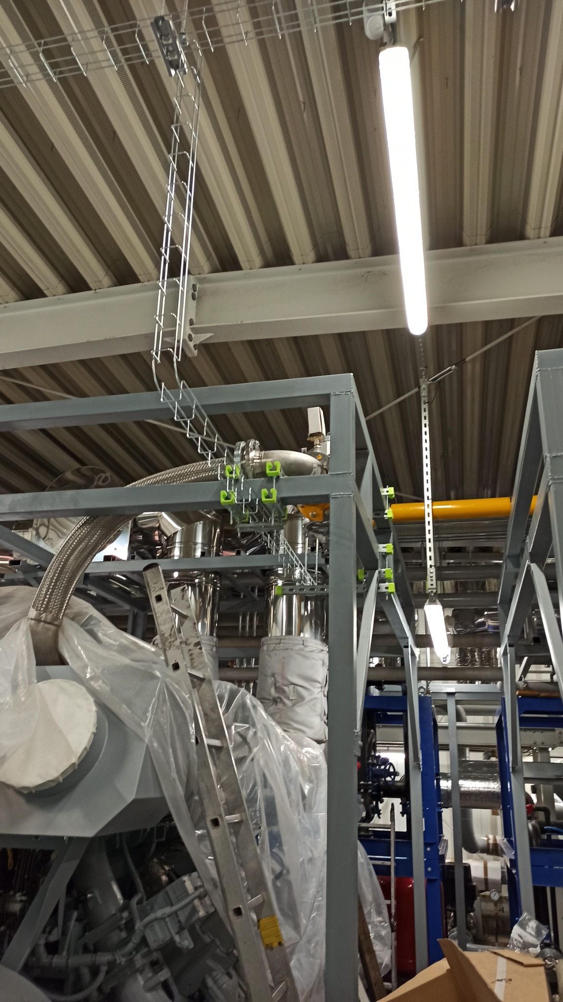 Widok na fragment hali produkcyjnej z instalacją przemysłową w trakcie montażu, z widocznymi elementami konstrukcji stalowej, rurami, przewodami w korytach kablowych oraz drabiną opartą...