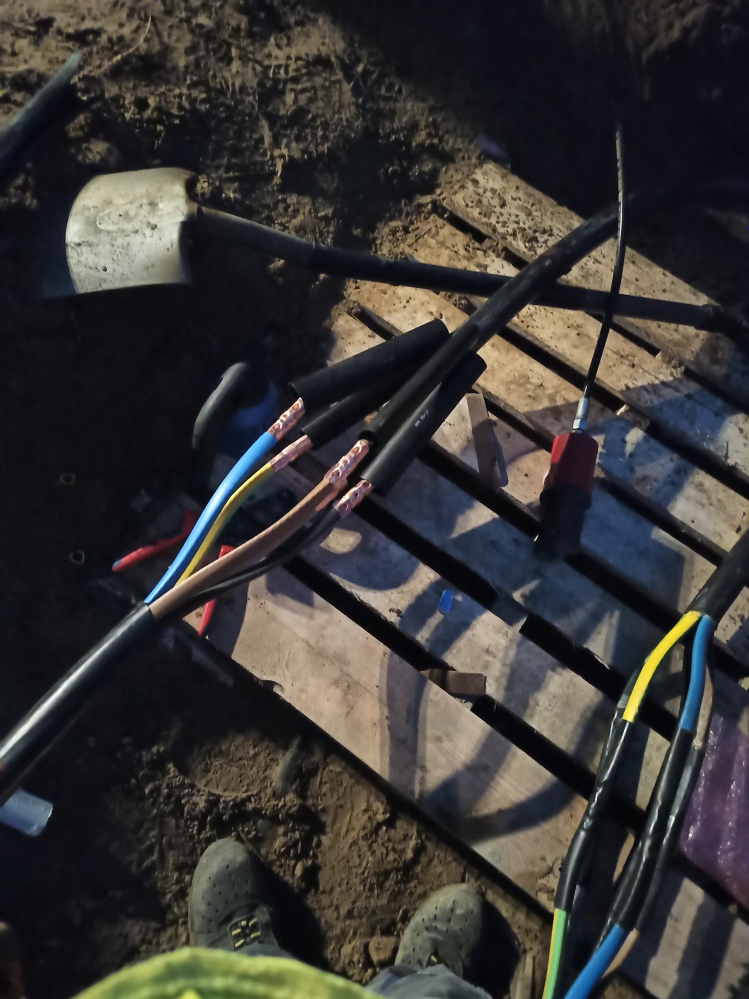 Fragment wykopu z ułożonymi przewodami elektrycznymi w czarnej izolacji, z widocznymi końcówkami kabli w kolorach niebieskim, żółtym i brązowym, przygotowanymi do połączenia, obok leży szpadel...