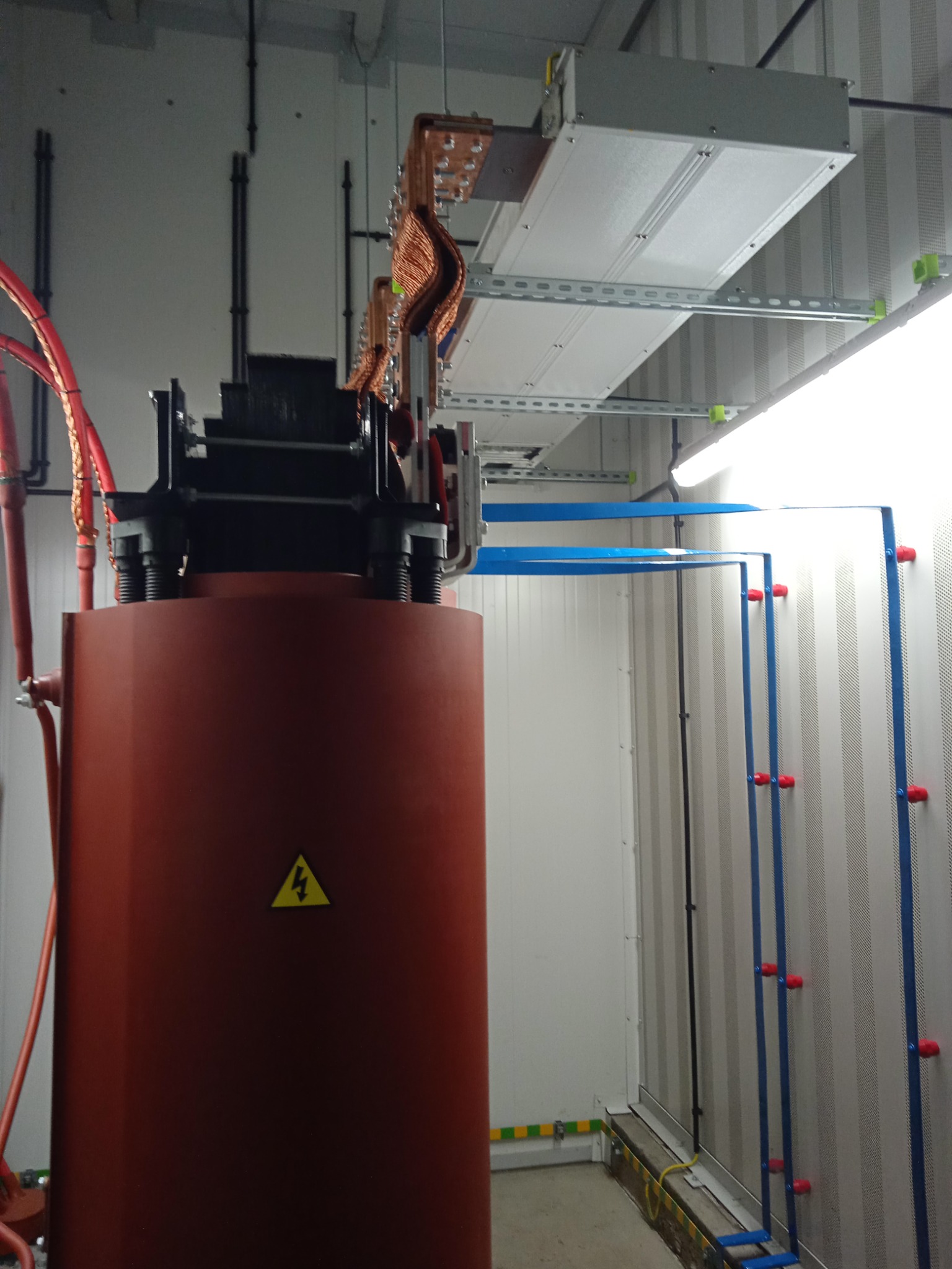 Brązowy transformator z żółtym znakiem ostrzegawczym na obudowie, widoczne miedziane przewody, szyny montażowe i oświetlenie w pomieszczeniu technicznym.