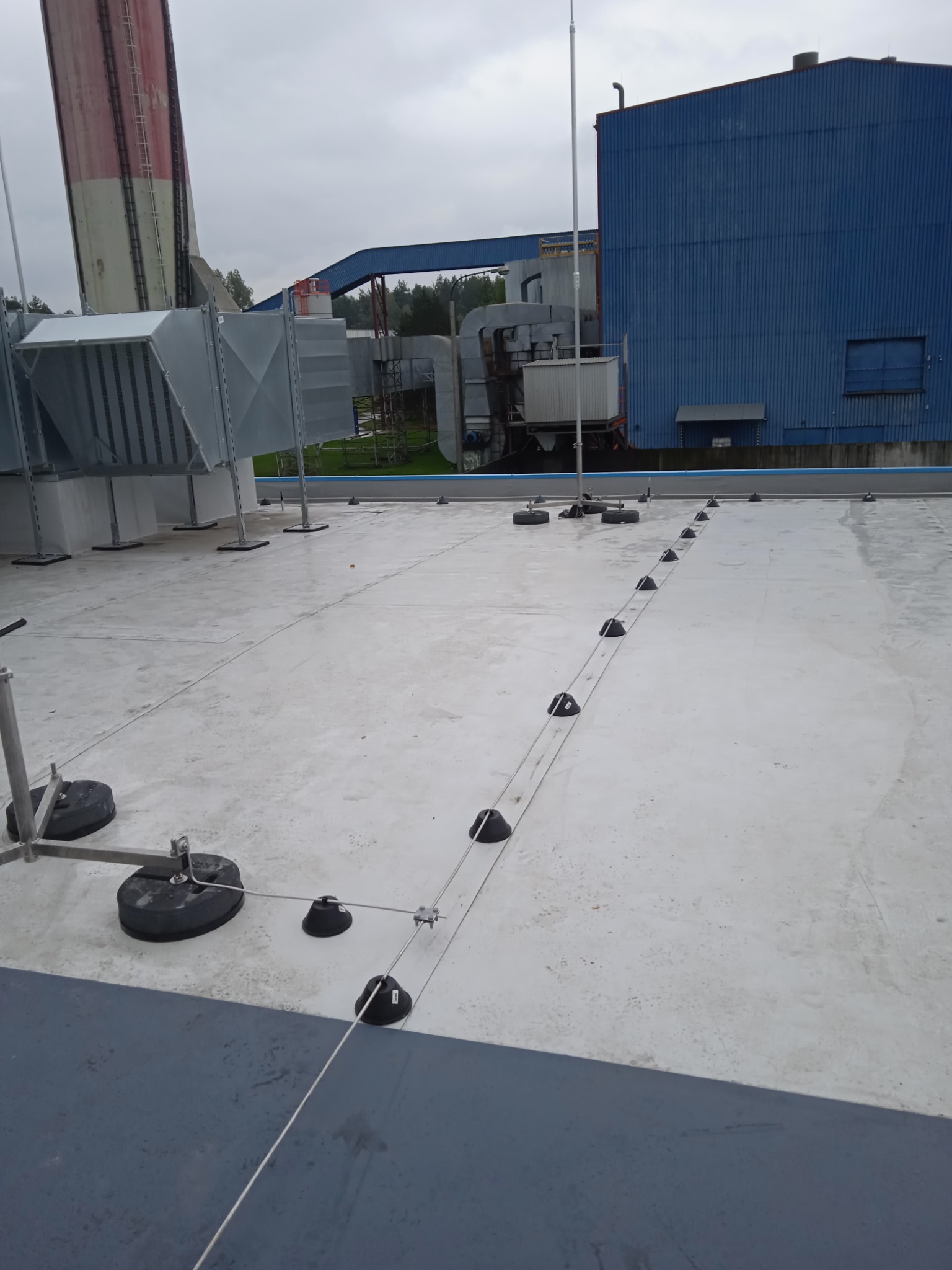 Instalacja odgromowa na dachu przemysłowym z widocznymi przewodami, uchwytami i masztem, na tle hali produkcyjnej i pochmurnego nieba.