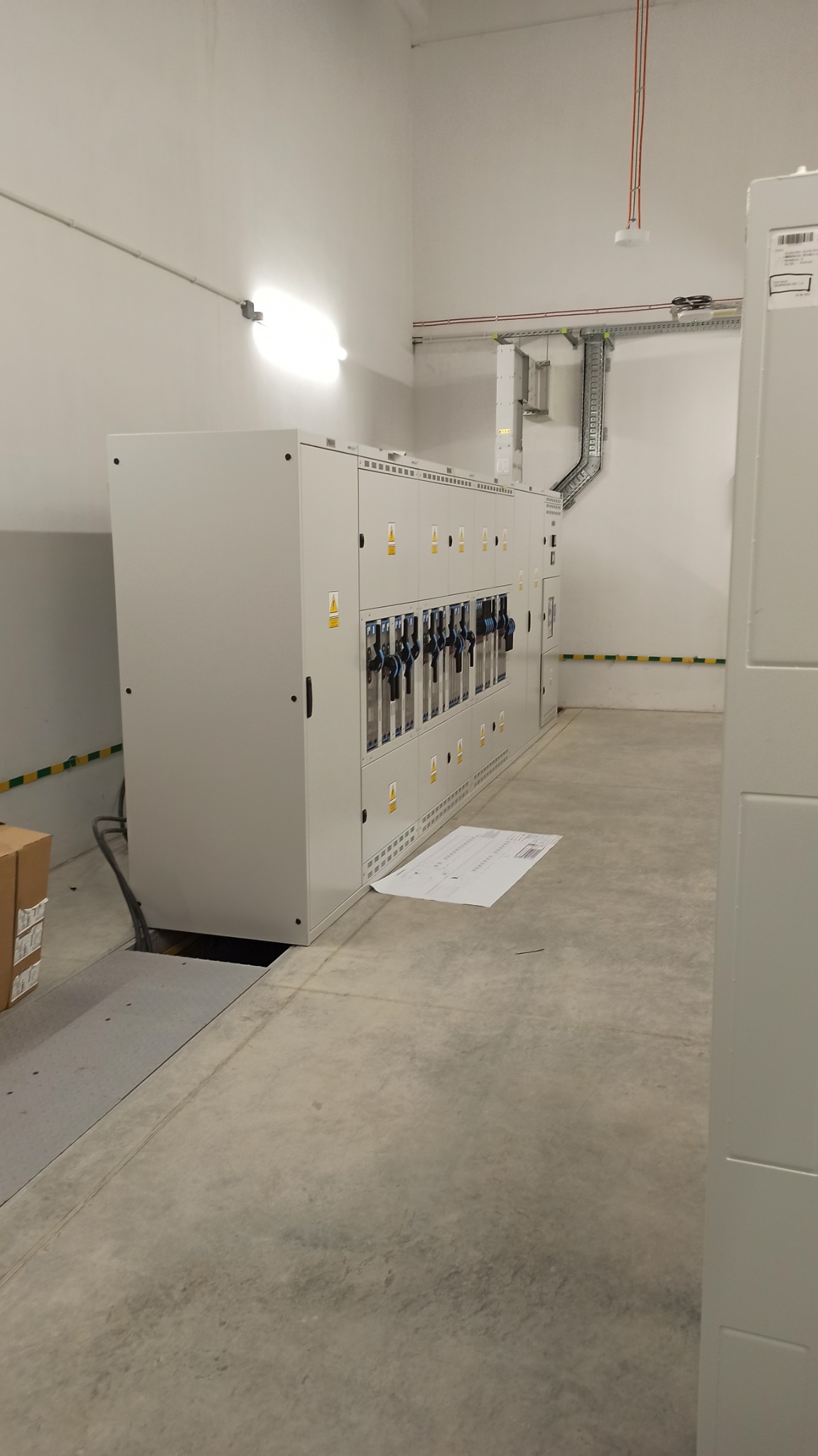 Rozległa szafa sterownicza z rzędami wyłączników i żółto-czarnymi oznaczeniami bezpieczeństwa w jasnym pomieszczeniu przemysłowym, z widocznymi kablami w korytach na ścianie i rozłożonym schematem...