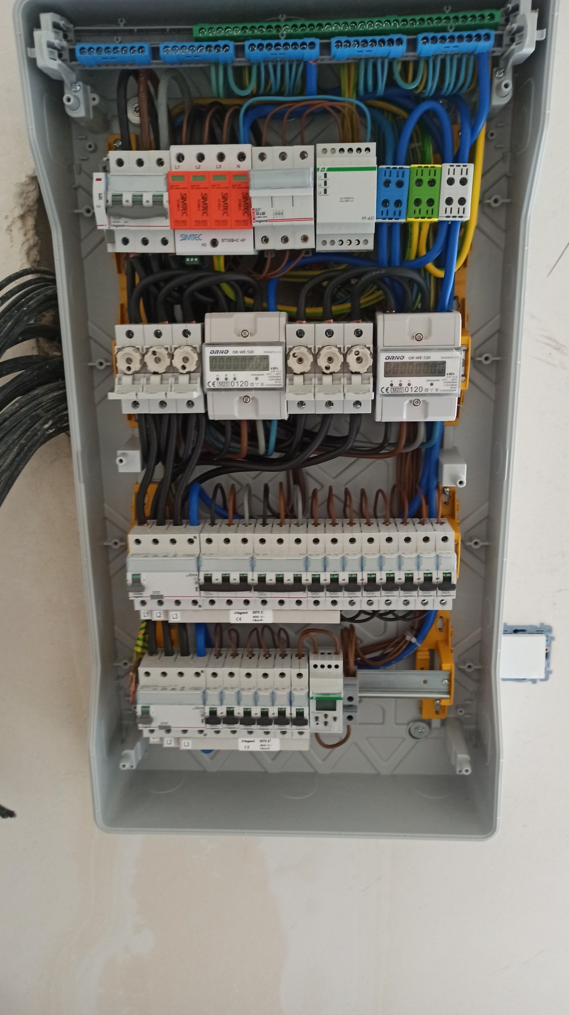Otwarta skrzynka rozdzielcza z zainstalowanymi bezpiecznikami, licznikami energii i okablowaniem, widoczne elementy firm Simtec i Orno.