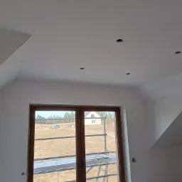 Poddasze ze świeżo pomalowanymi na biało ścianami i sufitem, widoczne otwory na oświetlenie punktowe i okno z widokiem na krajobraz.