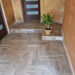 Korytarz z brązowymi płytkami imitującymi drewno ułożonymi w jodełkę, prowadzący do drzwi z przeszklonymi panelami, ściany wykończone w odcieniach beżu i brązu z dekoracyjną fakturą, doniczka...